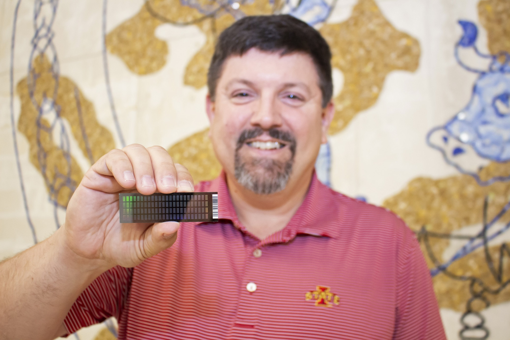 Smiling dark-haired man in red polo shirt holding computer chip, mural in background